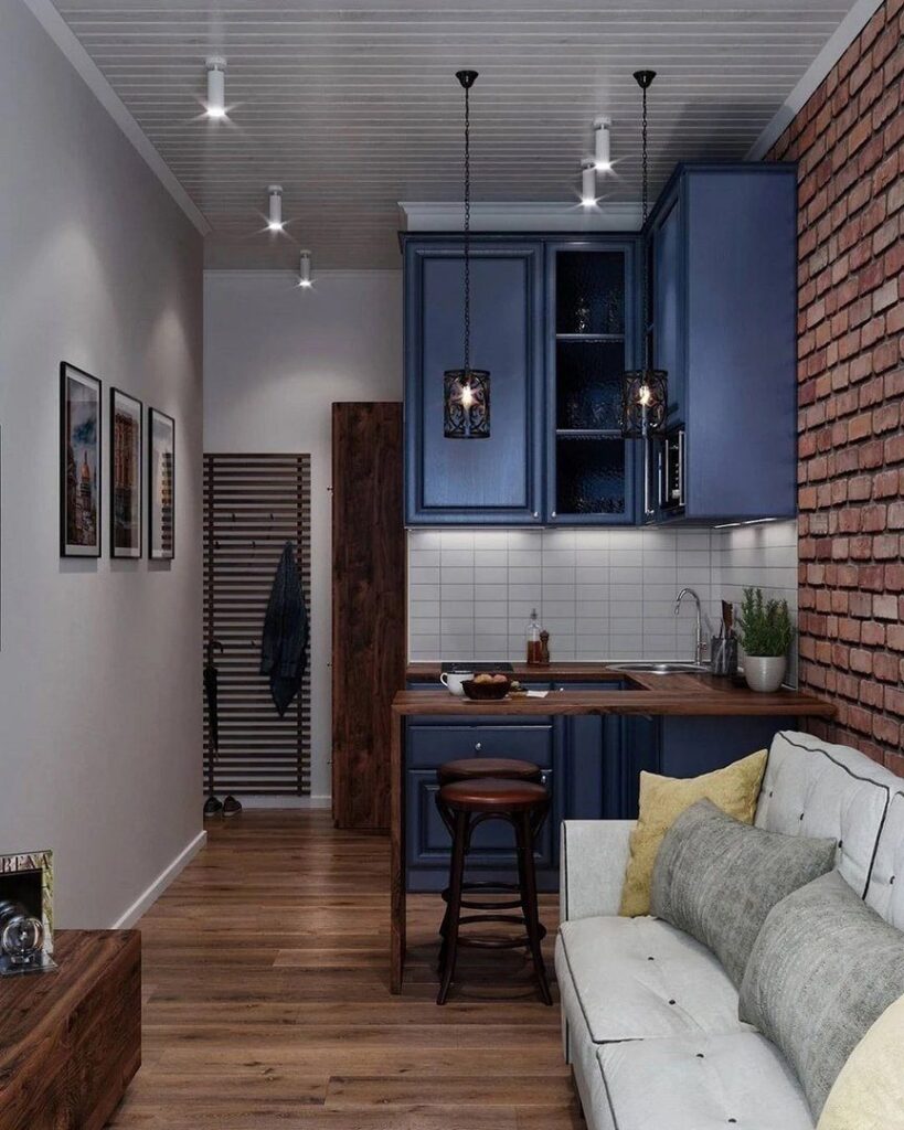 Small studio apartment interior with blue kitchen cabinets, white sofa, and brick wall.