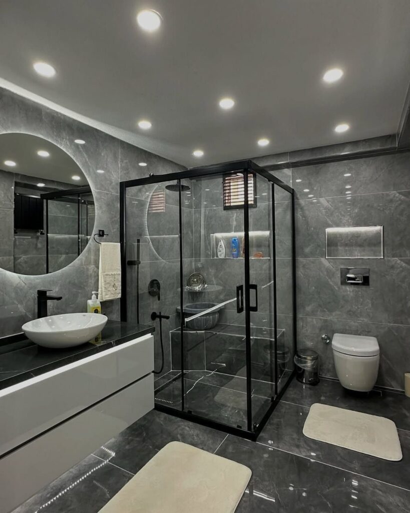 Large circular backlit mirror above a white floating vanity in a grey marble bathroom.