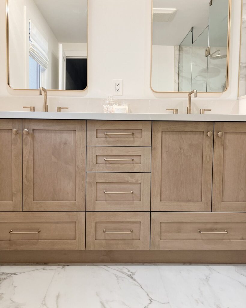 Modern bathroom vanity with light wood cabinets, gold hardware, and white marble floors.