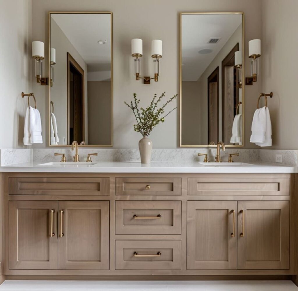 Double sink bathroom vanity with light wood cabinets, marble top, and gold mirrors.