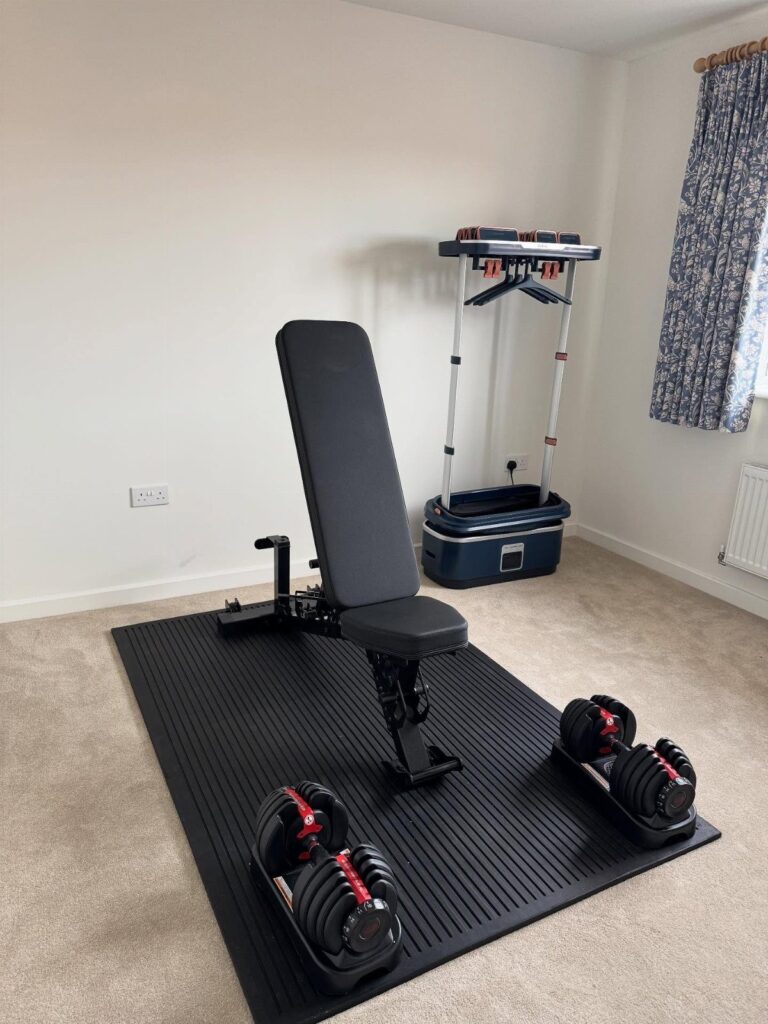 Weight bench and adjustable dumbbells on a rubber mat in a home gym.