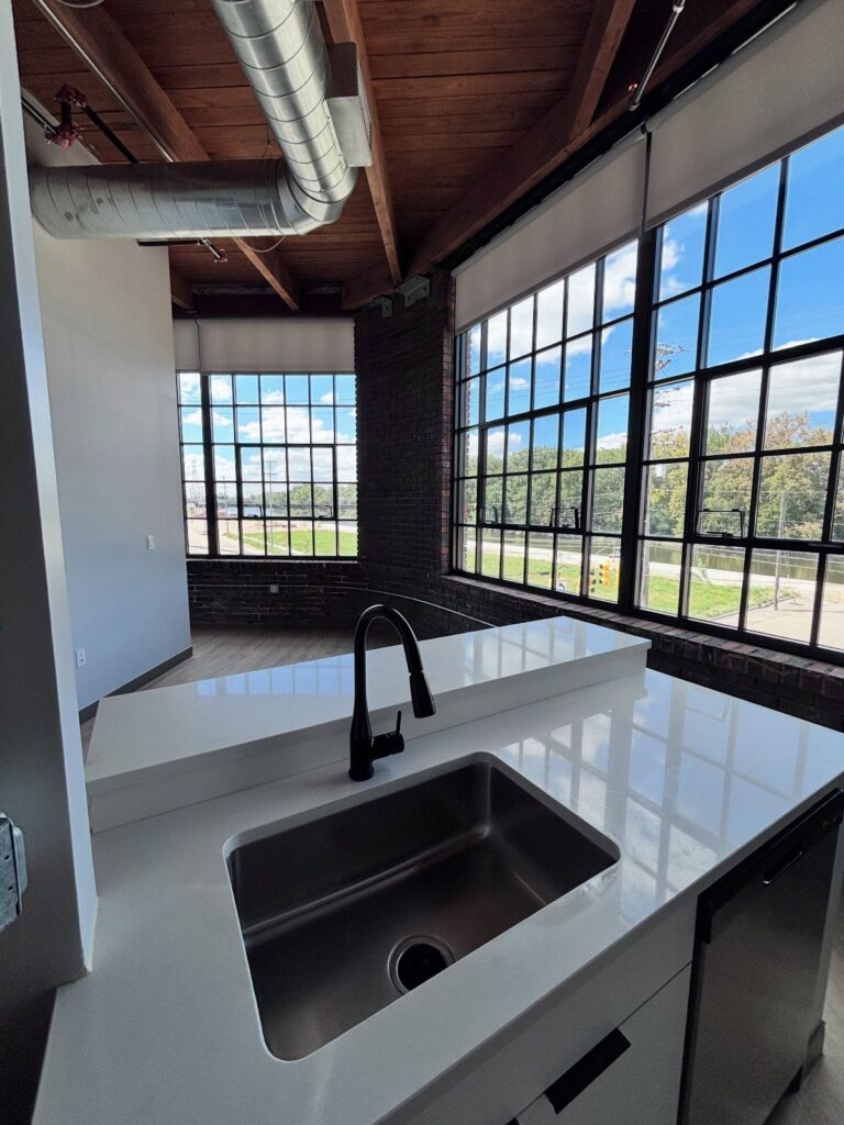 Modern loft kitchen with white countertops, exposed brick, and large industrial windows.