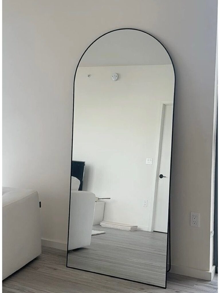 Arched full-length mirror with black frame leaning against a white wall in a room.