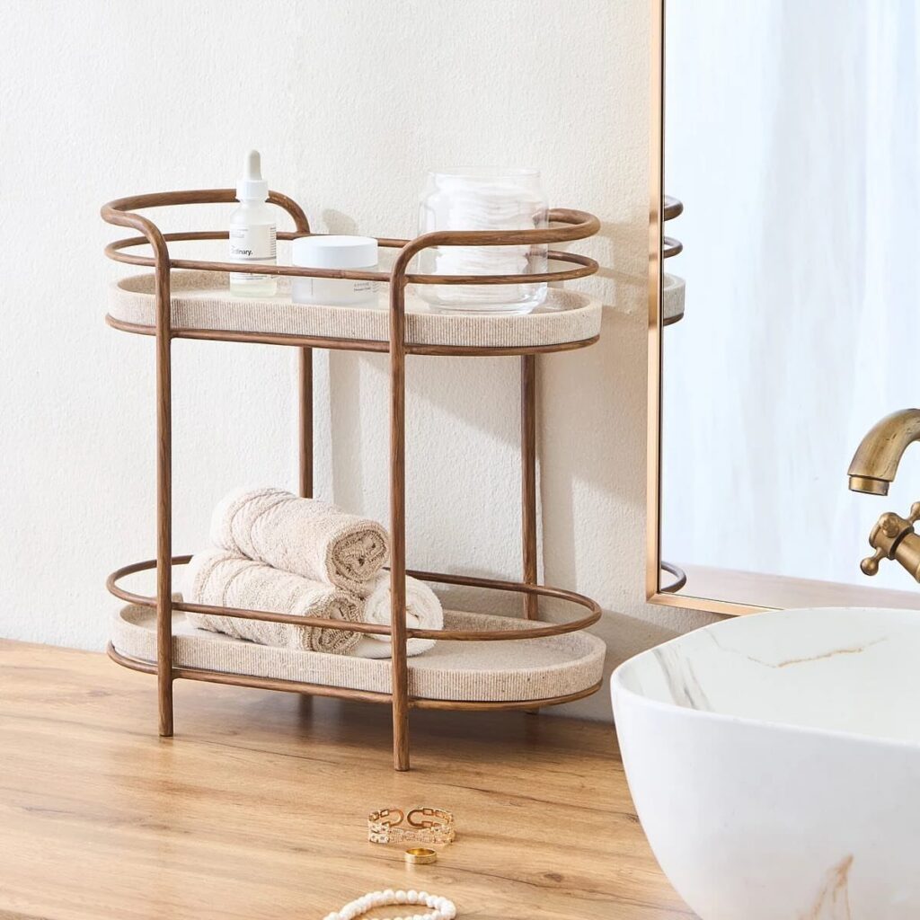 Two-tiered gold metal countertop organizer tray holding skincare products and rolled towels.