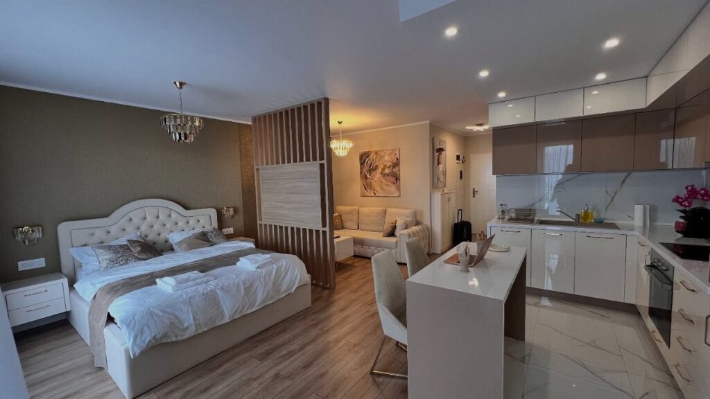 Modern studio apartment with a white bed, wooden divider, and integrated white kitchen.