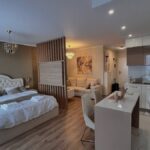 Modern studio apartment with a white bed, wooden divider, and integrated white kitchen.